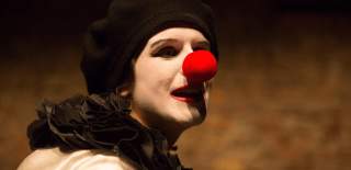 A person dressed as a clown, wearing white face makeup, a black beret, and a ruffled black collar, with a bright red clown nose. They are turned slightly to the side against a warm-toned background, with dramatic lighting highlighting their face.