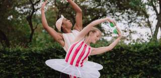Two people in ballet outfits wearing tutus. They are posing with their arms in the air over their heads holding green balls.