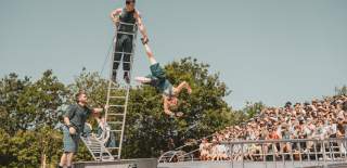 Three people doing acrobatics on a ladder on a stage. There is a crowd in seats to the right of the stage.