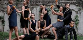 A group of eight adults pose theatrically in a garden against a brick wall, all dressed in dark formal or semi-formal outfits. Each person is holding oranges in stylised, almost sculptural poses, some standing, some seated, and one lying on the grass. One man crouches on all fours with an orange in his mouth while another person balances across his back. The group’s expressions are serious and composed, creating a surreal, staged tableau.