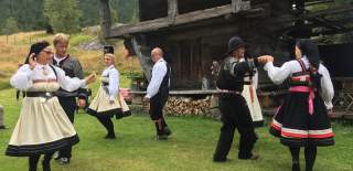 Menn og kvinner iført bunad danser ute på tun foran stabbur.