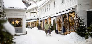 Snødekt gate med hvite trehus på hver side og julebelysning. En person leier sykkelen gjennom gaten