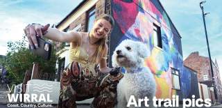 a girl taking a selfie with a white fluffy dog in front of a wall with a large art mural on it.