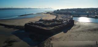 An aerial shot of a fort and castle on a beach in Wirral. To the left is the Mersey River and in the distance Liverpool skyline. To the right is a dock of water and build up Wirral.