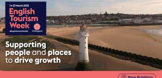 A lighthouse with a bright sandy beach, a bright blue sky and shops in the distance.