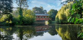A landscape shot of a beautiful still lake, surrounded by full, green trees and a red wooden walkway across the lake