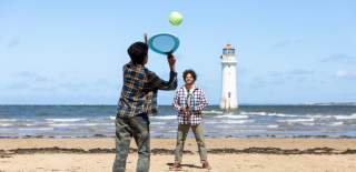 A family play tennis on the beach with New Brighton Lighthouse in the background