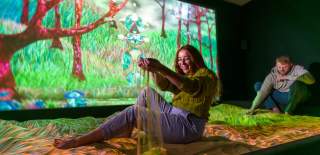 A lady sits in a sandpit in a dark room with a large, projected screen of nature behind her
