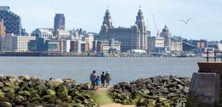 People stand on the shoreline surrounded by rocks looking over to the Liverpool skyline
