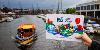 Hand holding a Love Bristol Gift Card shopping voucher from Pero's Bridge over looking the Harbourside, central Bristol - credit Bristol BIDs