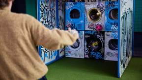 Person in a brown sweater standing on green turf, aiming a pink soccer ball at washing machines stacked in a graffiti-covered arcade game.