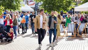 A busy outdoor street event in a sunlit city square, with two people walking through the centre holding drinks, surrounded by crowds, market stalls, deckchairs and trees.