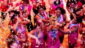 A women's netball team in pink uniforms celebrates with medals and a gold trophy amid falling confetti.