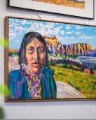 Photo of a painting featuring a native american looking into the eyes of the viewer from a colorful mountain background.
