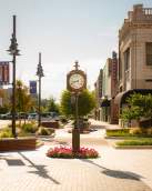 The City of Denison clock that is located in front of City Hall. It is a bright sunny day with clear blue sky. Pink flowers are in the bed around the base of the clock.