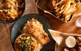 A top down view of a table filled with food at the George Tavern. The central plate has a piece of baked salmon on a bed of rice with a side bowl of Mexican street style corn. The bowl in the upper left corner is filled with a pasta dish. The upper right tray contains some chicken tenders and a pile of French fries.
