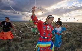 Women holding hoops surrounded by prairie hoop dancing in field under cloudy sky