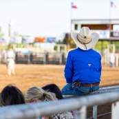 Fan looking on to the Start of the rodeo sitting on top of the railing in a cowboy hat.