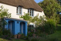 Charming cottage with a thatched roof and blue doors, surrounded by lush greenery and vibrant flowers, evokes a peaceful, idyllic countryside feel.