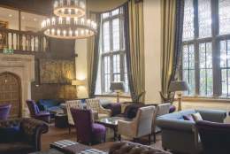 Luxurious room with plush velvet chairs, soft sofas, and a glass coffee table. A grand chandelier and tall windows create a warm, elegant ambience.