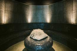 Dimly lit room with curved tiled seating, textured walls, and a glowing centrepiece of white crystals on a circular tiled pedestal. Calm, serene ambience.