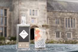 A bottle of handcrafted gin and a glass with a lemon slice sit on a table outside an old stone building, conveying a rustic and elegant vibe.
