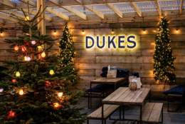 Festive outdoor seating area with wooden tables, benches, and Christmas lights. A lit sign reads "DUKES" on a wooden wall, conveying warmth and cheer.