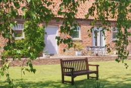 The outside of The Plovery at Church Farm Cottages in East Yorkshire. A wooden bench sits in a shaded area, under the hanging branches of a large tree.