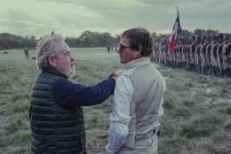 Actors stand in a field with soldiers in thr background