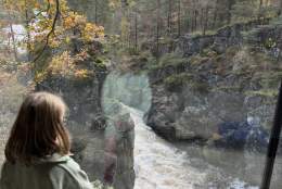 Et barn sitter ved et stort vindu og ser ut på Kvåsfossen i Lyngdal. Elva fosser ned gjennom kløften omgitt av høstfargede trær.