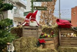 En juledekorert scene i sentrum av Lyngdal, med halmballer, trekasser fylt med julepynt, to nisser, røde julestjerner og et grønt veiviserskilt som peker til lokale attraksjoner. Små lyslenker og et tre i bakgrunnen gir førjulsstemning.