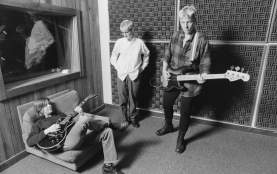 Photo of Meg Hentges and Darcee Douglas and another musician in a studio Douglas is standing on the right and playing the guitar, Hentges is standing in the middle and looking down and a third musician is sitting on a cushion on the left and playing the guitar
