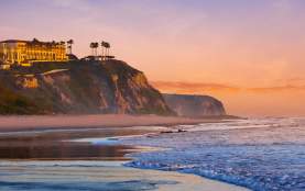 Ocean waves wash onto a Dana Point sandy beach at sunset with cliffs and yellow buildings topped with palm trees in the background.