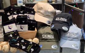 Socks with "Gilroy, California" and whimsical garlic bulbs, and Ball Caps of different colors with "Gilroy, California" displayed on a shelf.
