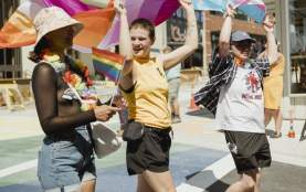 Three people walking on the street, each holding a different flag over their heads. From left to right- Pride flag, lesbian flag, and trans flag.