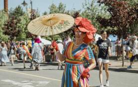 A person holding a parasol dressed in rainbow with a large flower hat posing for a photo.