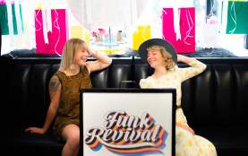Two smiling women in vintage outfits sit on a black leather couch inside Funk Revival, with colourful shopping bags and decorations displayed behind them.