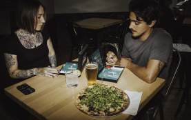 Two people play a tabletop game at a restaurant table with pizza and drinks in front of them.