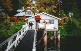 Two people stand on a narrow walkway leading to a small oceanfront house on stilts above the water, surrounded by trees and fall foliage.