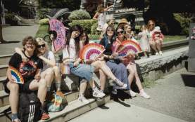 A group of 6 people holding a variety of pride themed fans, smiling and embracing each other.