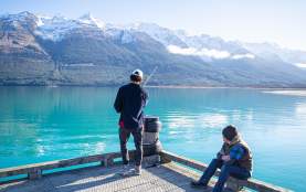 A man fishing off a wharf over a vibrant blue lake and snow covered mountains, while another sits and watches.