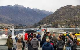 A group of people listening to a person doing a speech on Queenstown Marina with an electric boat and The Remarkable Mountains in the background