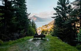 Two people sitting on a chair at the top of a lookout with tall trees in foreground and lake and mountain in background