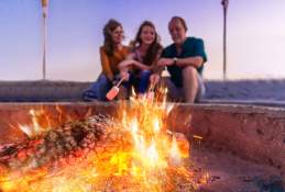 A family of three sitting by a bonfire on the sand roasting marshmallows