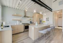 A modern kitchen, with light wooden cabinets, island, and floor and stainless steel appliances. The wall is a very light turquoise.