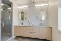 A modern-style bathroom with a large shower on the left hand side with a glass door, a light brown wooden vanity, and two large round gold mirrors hanging over the two sinks.