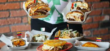 A person offering the viewer tortas in both hands with a table of tortas, empanadas and more food below