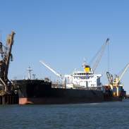 An Iron ore carrier in the Port of Port Hedland, seen from the water with a tug boat along side and a crane on land.