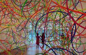 Two people can be seen in the distance through a piece of artwork that has multicolor strands arranged in circles.