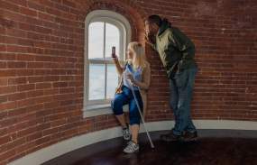 A man and woman are seated inside a room of the Tarrytown lighthouse. The man is looking out the window while the woman, seated on the windowsill, is taking a picture of the outdoor view.